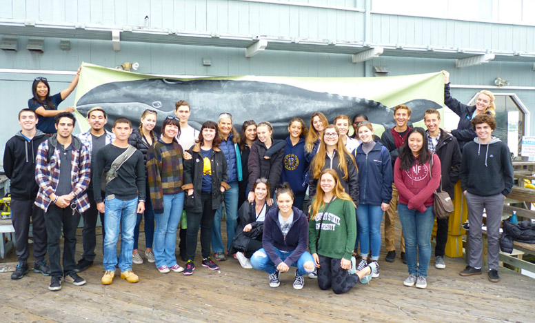 A group of children and young adults pose for a picture with marine science educators in front of a banner of a whale.